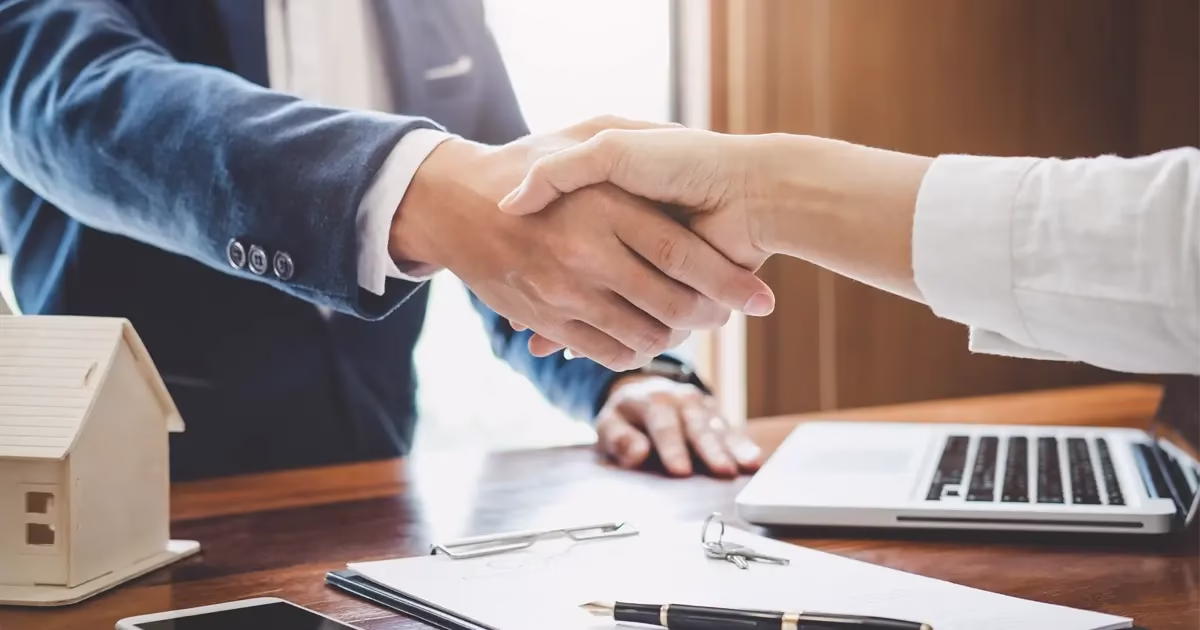 Deux personnes se serrant la main au-dessus d'un bureau avec des documents, un ordinateur portable et une maquette de maison